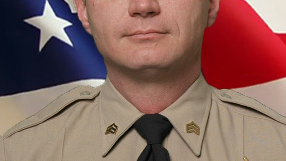 Law enforcement officer in tan uniform with sheriff's badge, standing before American flag