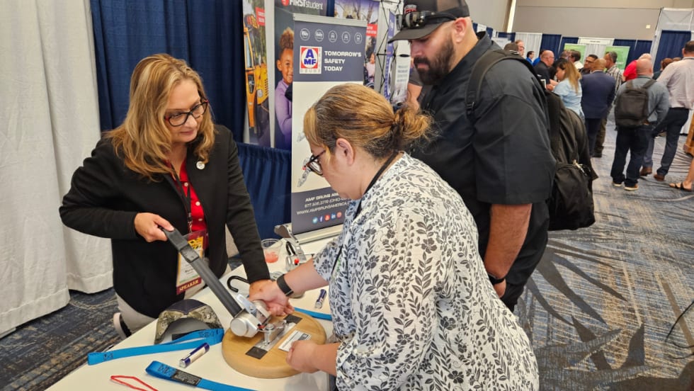 Attendees visit the AMF Bruns vendor booth in the expo hall, examining a wheelchair securement device while speaking with an exhibitor about safety equipment.