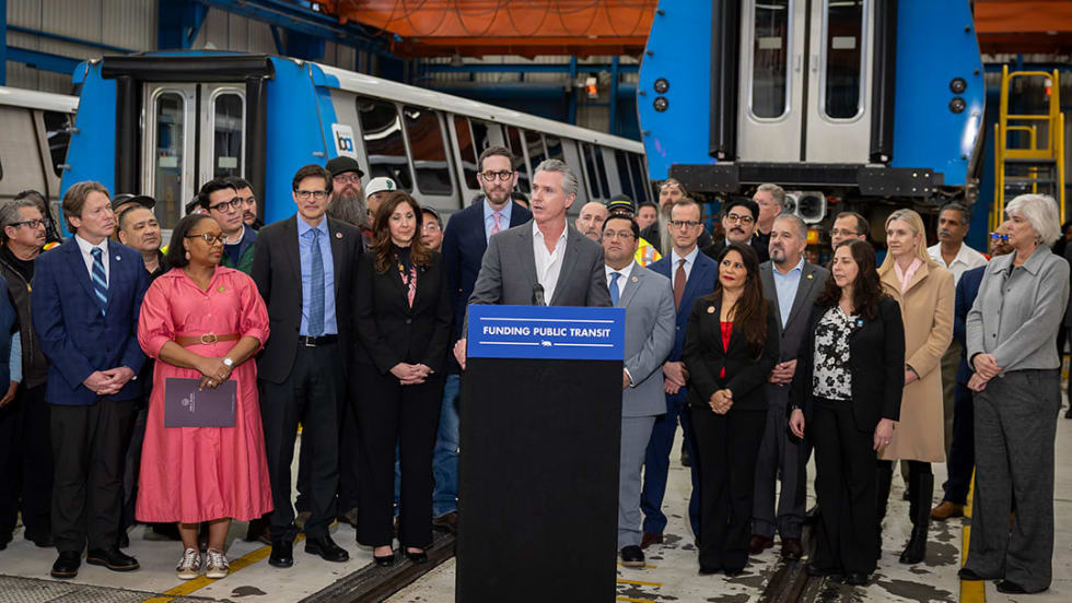 California Governor Gavin Newsom stands at a podium surrounded by a group of state and community leaders.