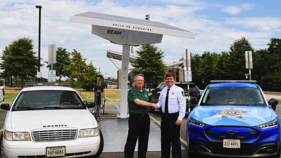 A white Ford Crown Victoria Police Interceptor and blue Mustang Mach-E are shown sitting in front of a Beam solar charger. The fleet manager and sheriff are seen shaking hands between the vehicles.