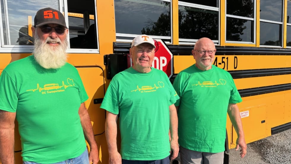 Three men in green shirts pose next to a school bus.