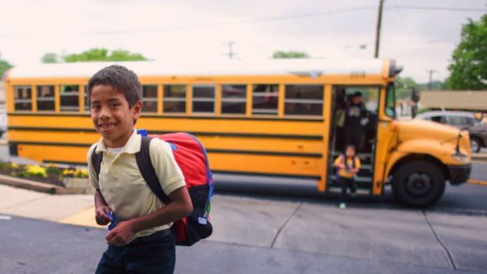 a child walks on the sidewalk with a school bus behind him