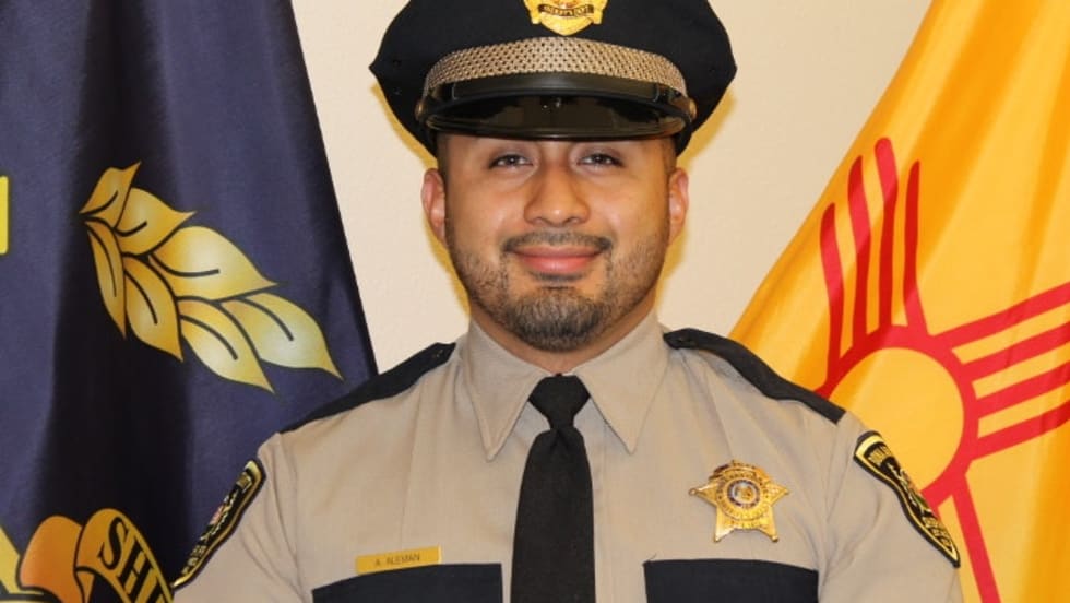 Sheriff's deputy in tan uniform and black cap, standing in front of state flags