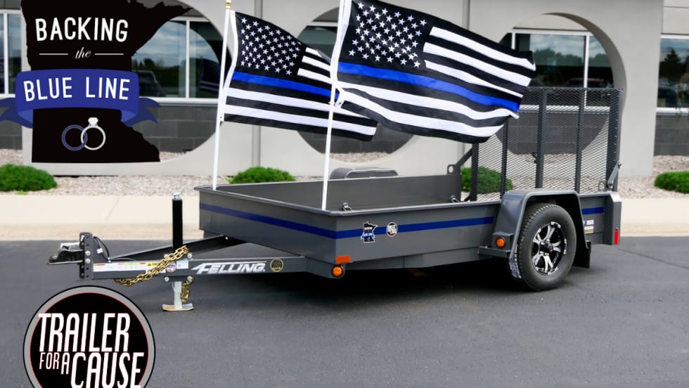 A dark gray dropdeck utility trailer with a thin blue line is shown, with 2 Thin Blue Line flags hanging off the back.
