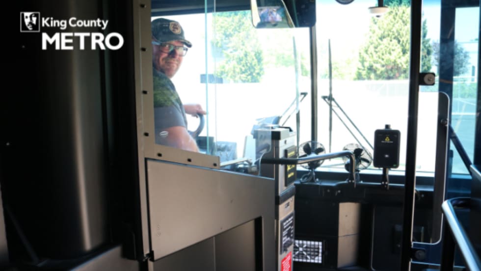 Image of a bus operator posing from the front seat of a public bus, behind a clear partition.