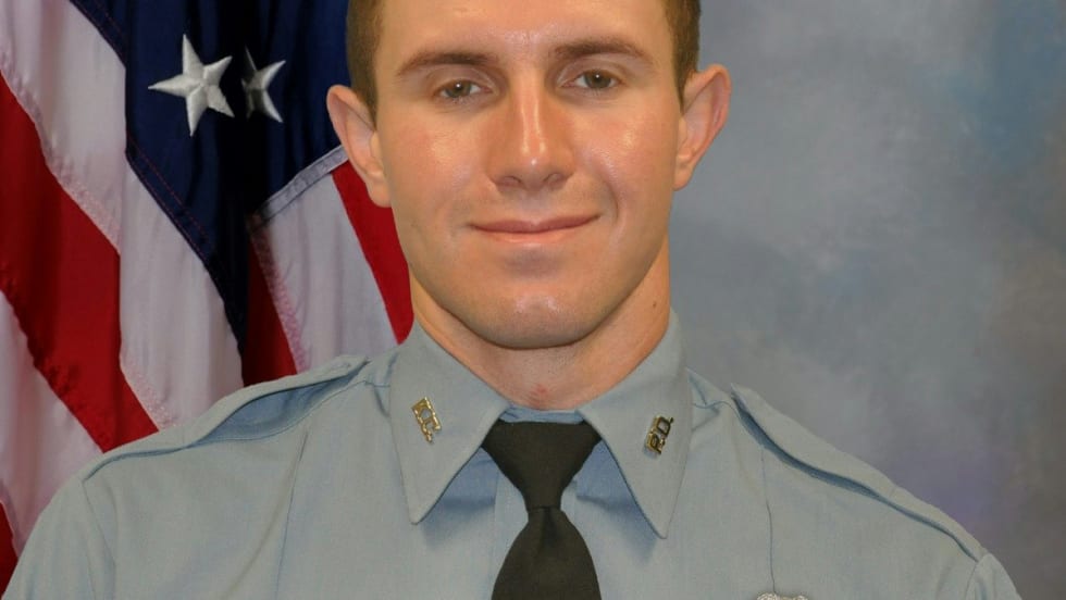 Police officer in light blue uniform standing in front of American flag