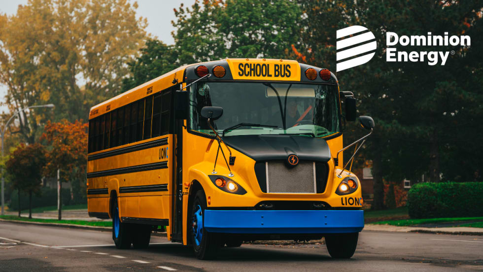 A LionC all-electric school bus drivers down a road with colored leaves in the background.