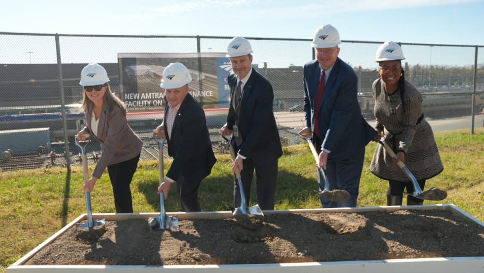 Five people in business attire lined up shoveling dirt.