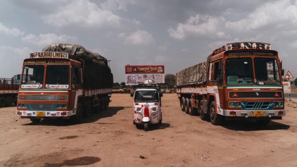 A rickshaw parked between two red transit buses in India.