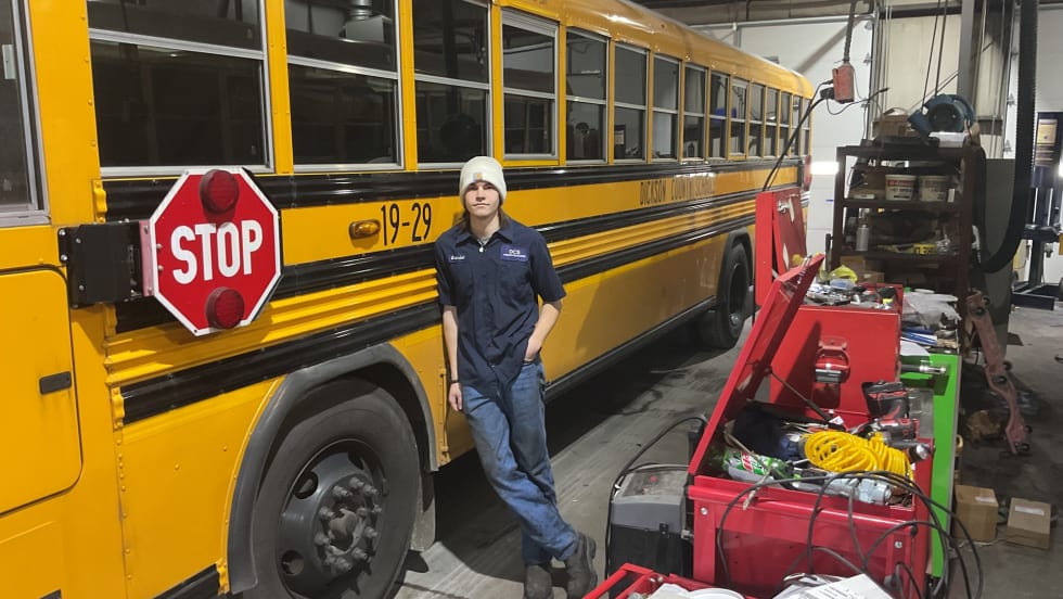 a young man leans against a school bus in a maintenance garage