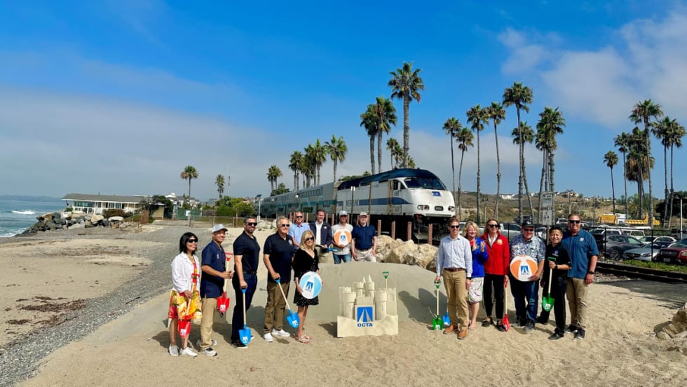 A group of people stand on a beach with a rail car in the background.