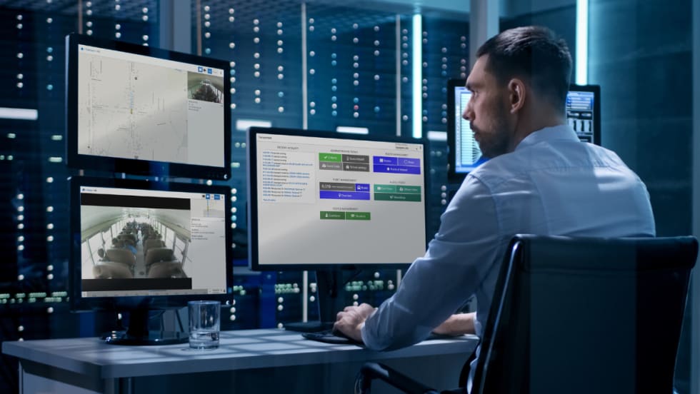 A man sits at a desk with three computer monitors displaying Transportant's platform.