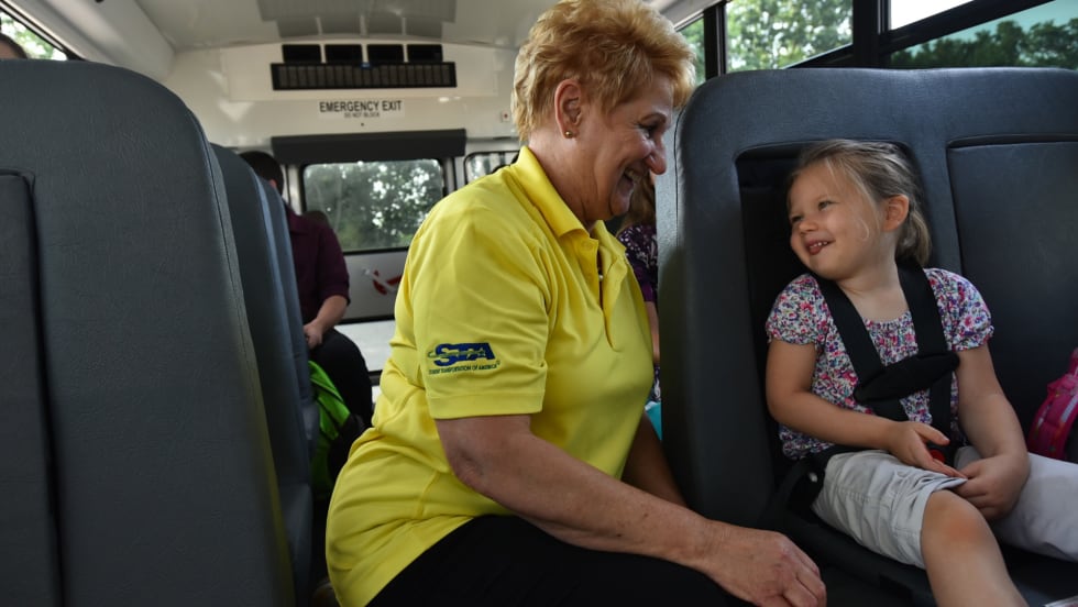 A school bus driver from Student Transportation of America smiles and talks with a young girl buckled safely into her seat on the bus.