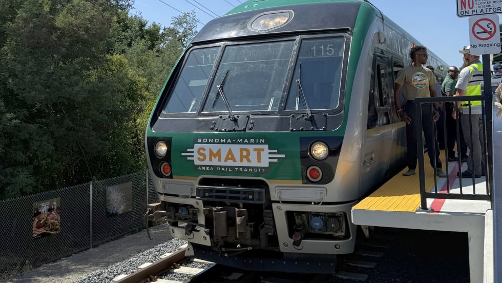 A SMART (Sonoma-Marin Area Rail Transit) train stopped at a platform during a station opening event, with people standing beside the train and signage posted near the edge of the platform.