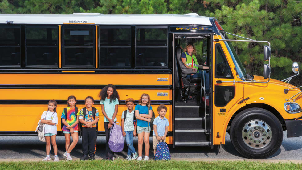 A group of students stands in front of a Thomas Built Jouley electric school bus. The door is open with the bus driver smiling for the camera.