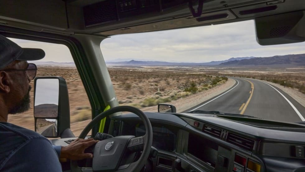 Truck driver navigating a winding desert highway, viewed from inside the cab of a Volvo semi-truck, with mountains and open landscape visible through the windshield.
