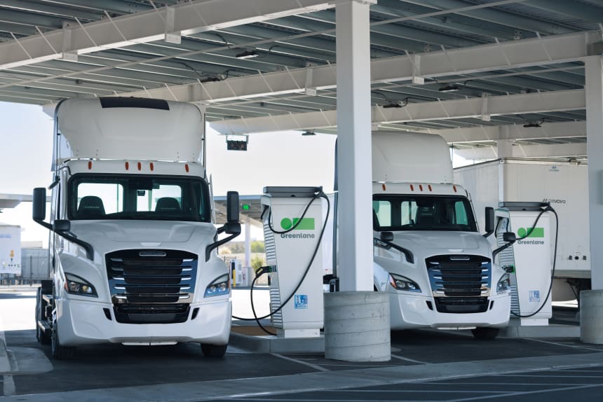 Two white electric semi-trucks at a Greelane charging station under a covered depot, showcasing sustainable fleet technology and EV freight infrastructure with charging stations.
