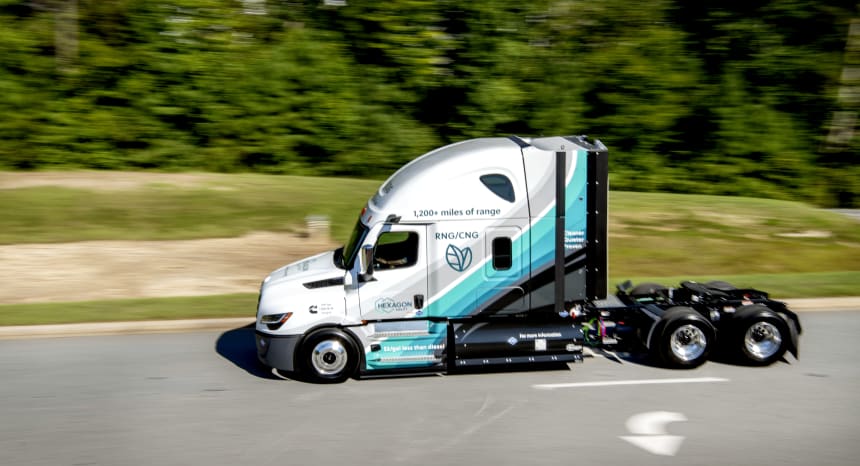 Aerodynamic teal-and-white semi truck cab races down a highway past forest, motion-blurred background highlighting sleek commercial transport. Driver's side door has the Hexagon Agility logo. Upper Cab has RNG/CNG with leaf graphic and "1,200+ miles of range" statement.
