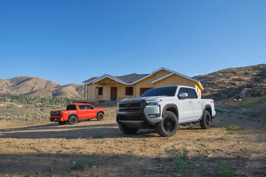 Two Nissan pickup trucks, one white and one red, are parked in a dusty, semi-arid landscape in front of a house under construction.