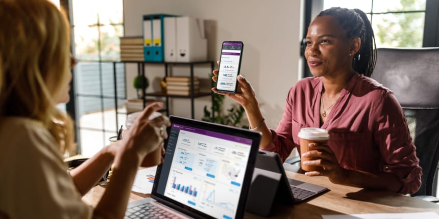 Two colleagues collaborate in a bright office: a woman demos the PrePass mobile app while another reviews analytics on a laptop, coffee and charts nearby.