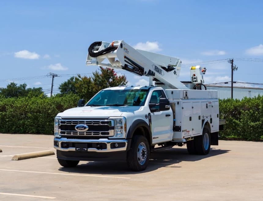 White Ford utility truck with an extended bucket lift arm parked in an empty asphalt lot with green bushes and a building in the background under a bright blue sky with scattered clouds.