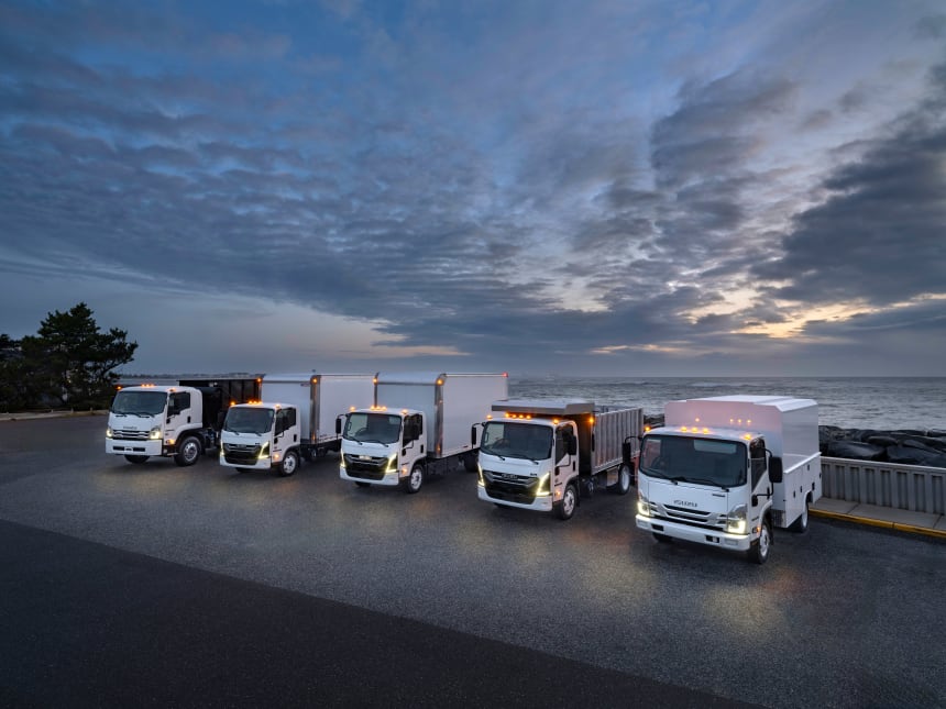 Isuzu commercial fleet of N-series and F-series white trucks lined along a coastal road at dusk, headlights glowing under dramatic clouds—reliable fleet service by the ocean.