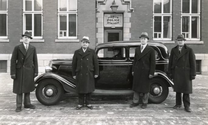 Four police officers standing beside vintage police car in front of station