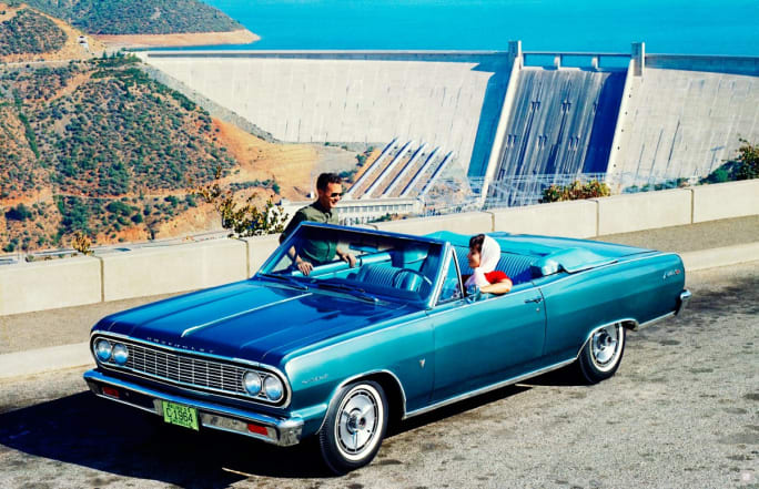1960's blue convertible parked on the side of the road with a woman inside and man outside. A large dam is in the background.
