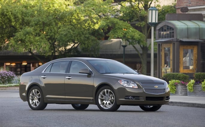 Gray car parked on a city street with buildings and trees in background.