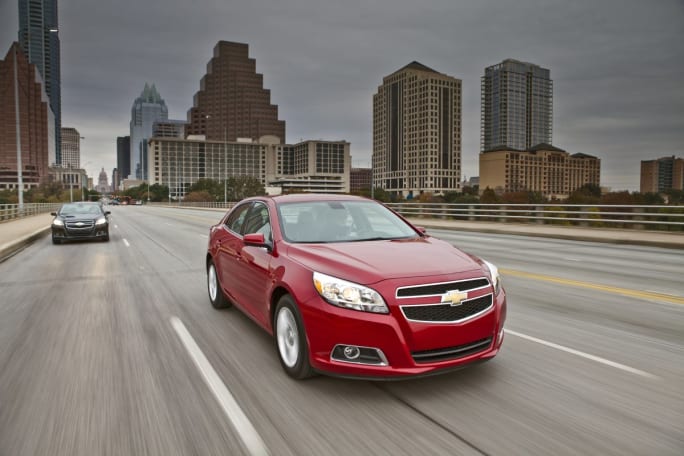 Red car drives down multi-lane road with tall city buildings in background.