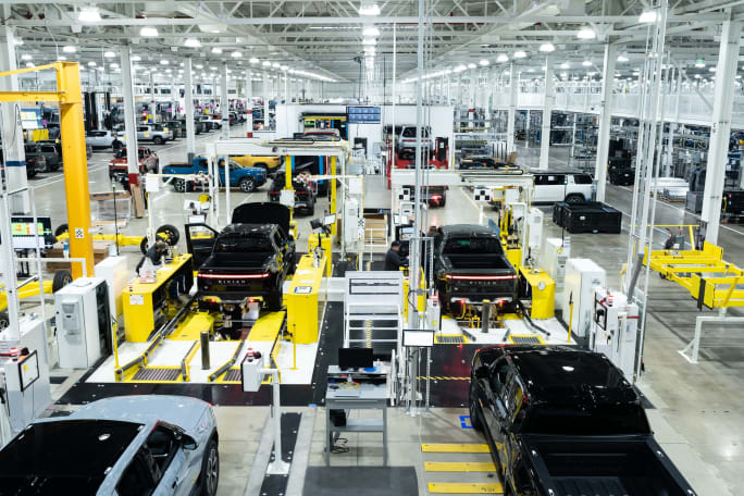 Rivian vehicle assembly line inside a large manufacturing plant with multiple electric trucks being built.
