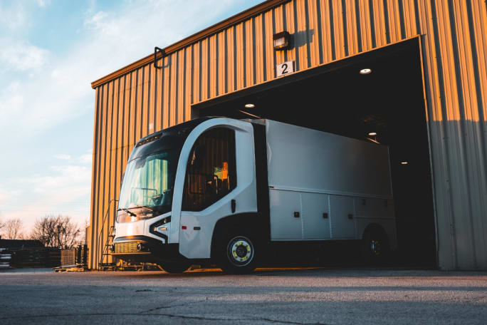 A REE P7-C truck drives out from inside a garage or shop building.