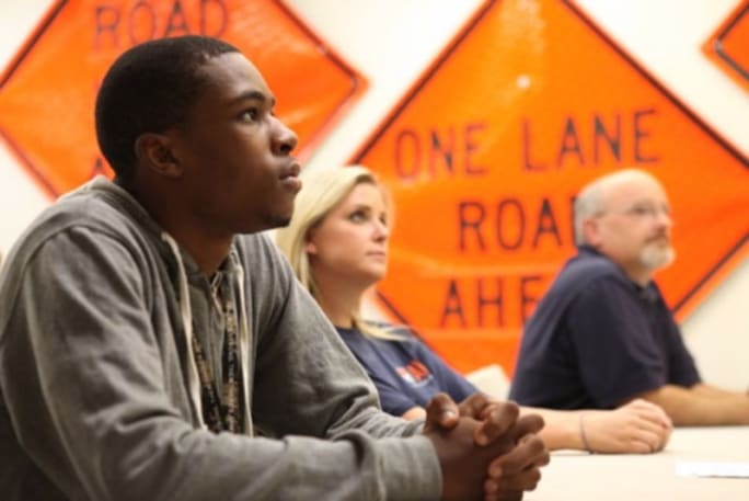 Workers seated at tables with orange construction safety signs in the background.