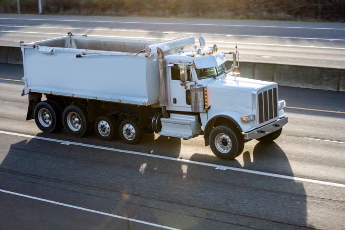 White dump truck driving on highway from left to right, with two extra axles underneath.