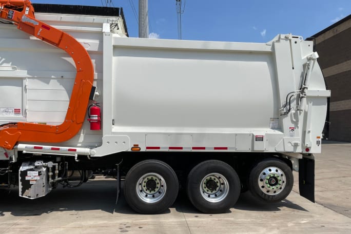 Side photo of the rear of a white refuse truck, facing left, with a lift axle installed near the rear.