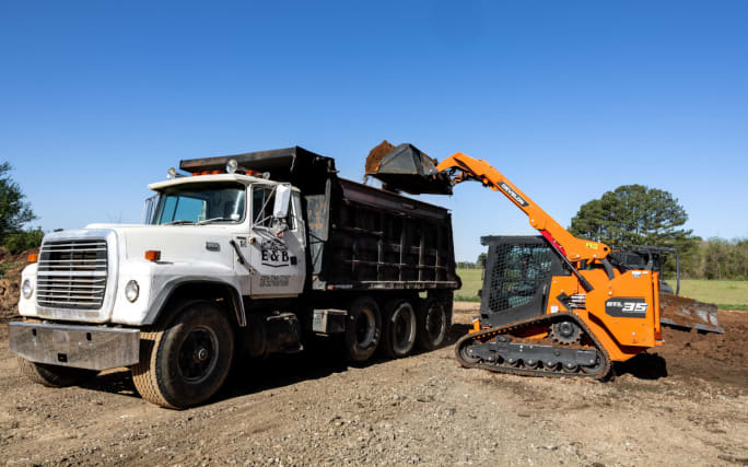 An orange compact loader placing dirt into a dump truck.