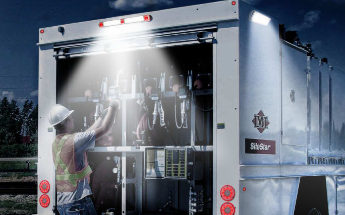 Worker at tool storage area at rear of work truck, illuminated by overhead light.