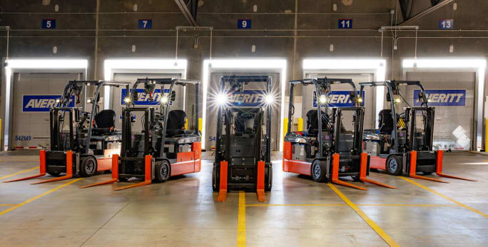 Line of electric forklifts with lights on in Averitt cross-dock with trailers in loading doors behind them.