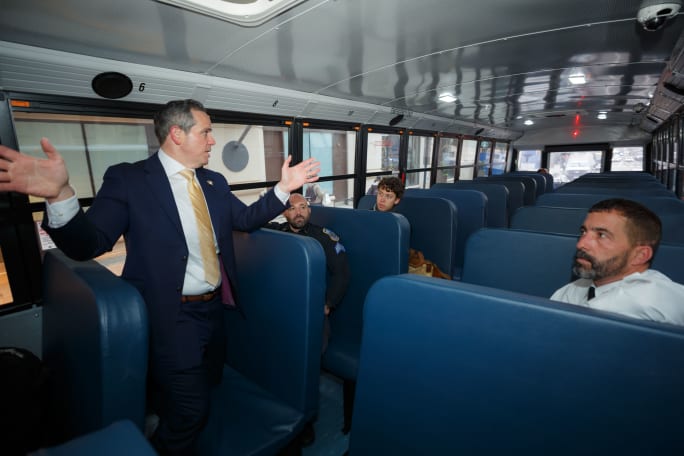 A man in a suit speaks to people inside a school bus.