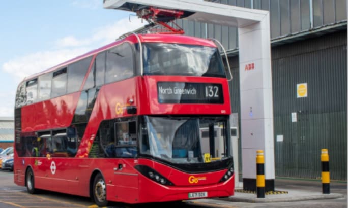 A red electric double-decker bus in London charges at a fast-charging station, with its pantograph connected above the bus while displaying its route to North Greenwich.