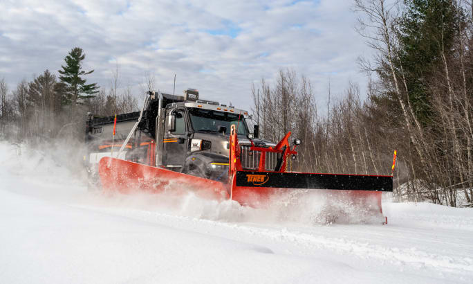 Western Star 47X snow plow truck from Alamo Group clears snow on a rural road, with trees in the background and snow flying off the blade.