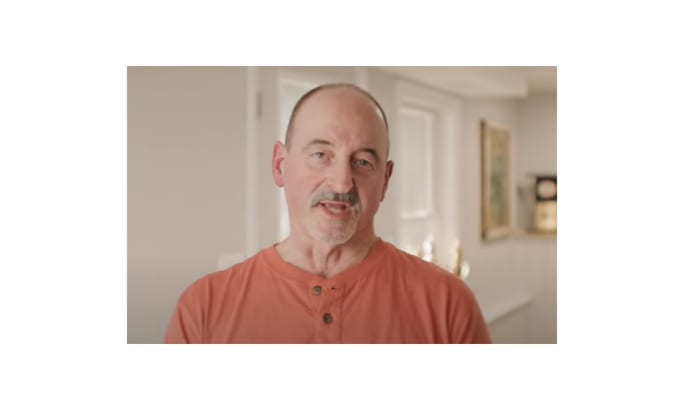 A man in an orange shirt speaks while standing indoors, featured in the ATA’s DRIVEN series highlighting the stories of professional truck drivers in the United States.