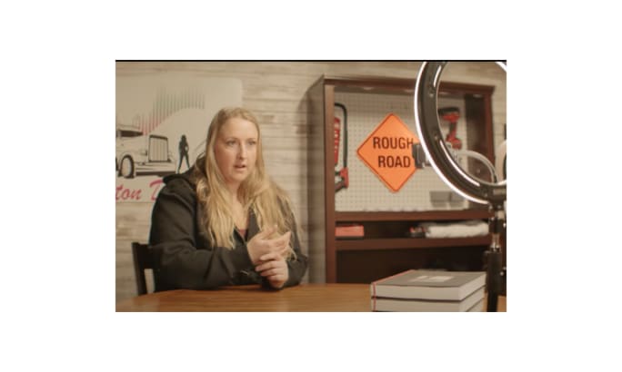 A woman with long blonde hair speaks while seated at a table, with a “Rough Road” sign and truck-themed decor behind her, featured in the ATA’s DRIVEN series on professional truck drivers.