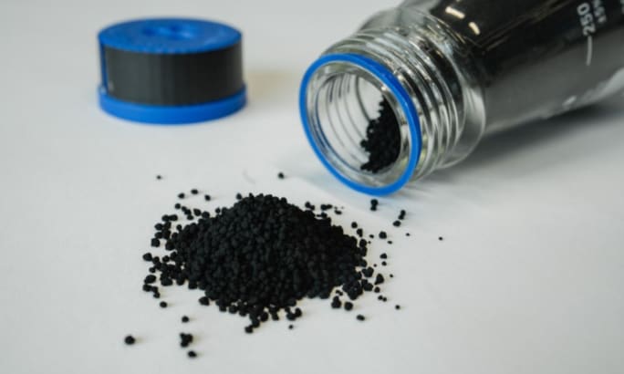 A small pile of black granular material spilled from a clear lab jar with a blue rim, lying on its side on a white surface. The blue cap is placed nearby in the background.