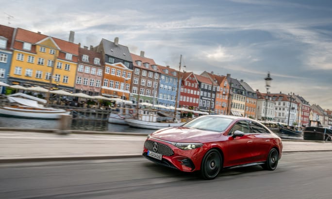 A red Mercedes-Benz CLA drives alongside a canal lined with colorful historic buildings, capturing the vehicle in motion within an urban European setting.