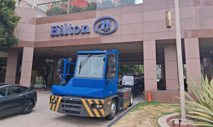 Blue ZM Trucks T75 electric terminal tractor displayed outside a Hilton hotel entrance.