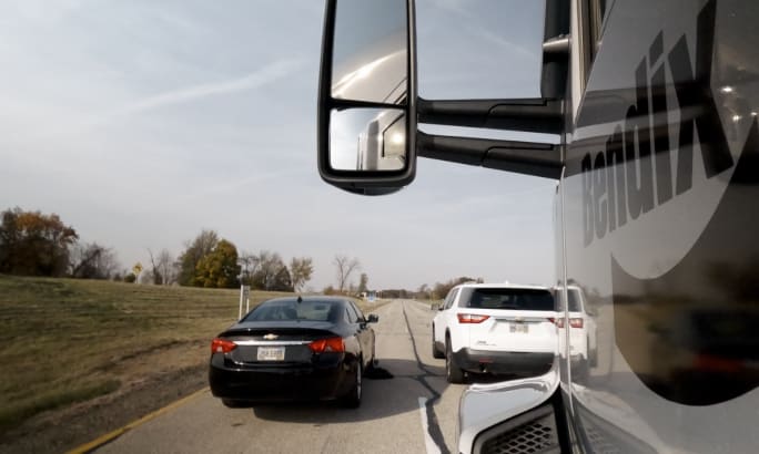 Side mirror view from a Bendix truck showing nearby black and white vehicles on a highway.