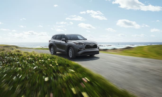 Gray Toyota Highlander SUV driving on coastal road with green grass and ocean background.