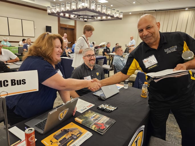 A man and a woman shakes hands across a table in a large room during an event.