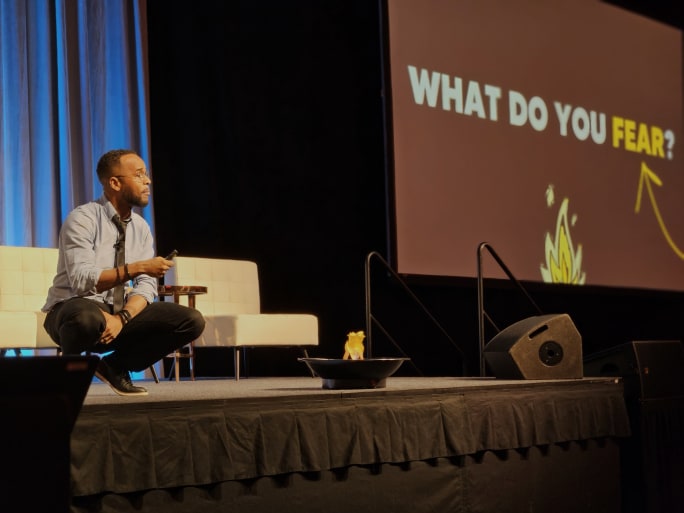 a man crouches on a stage with a screen behind him that reads 